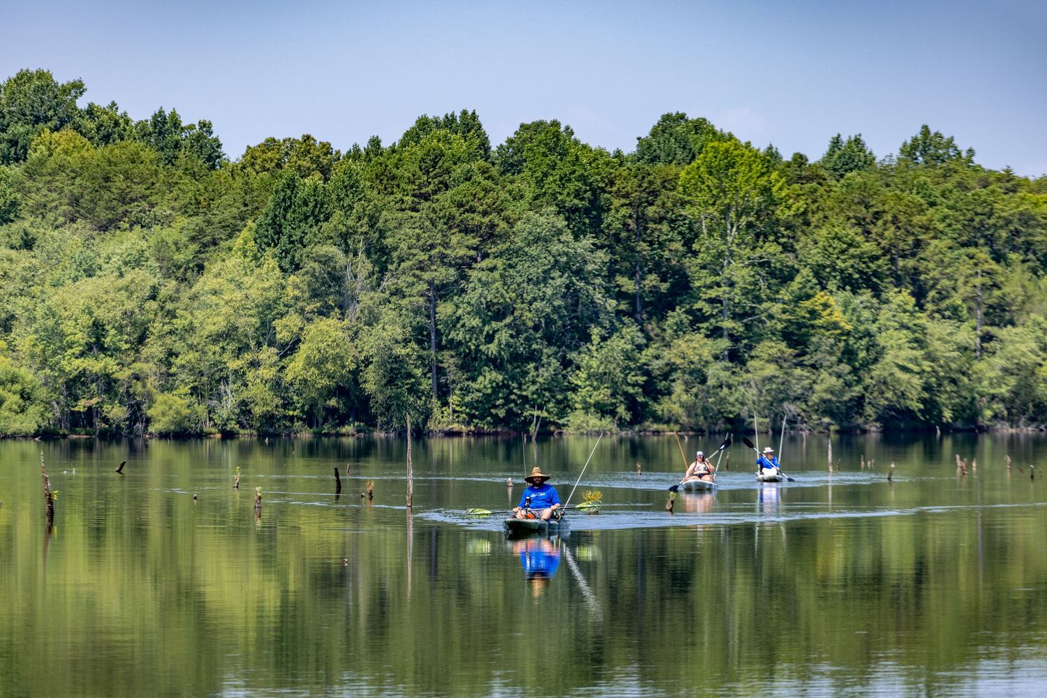 Kayakers paddling on a calm reflective lake surrounded by lush green trees on a clear summer day.