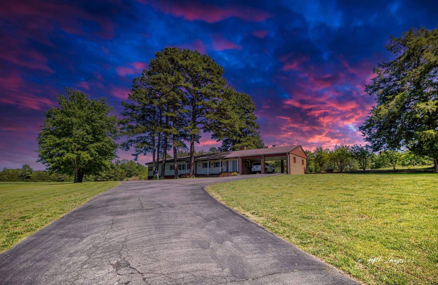 Ranch-style brick home with long driveway, tall pine trees, and vibrant pink and purple sunset sky backdrop.