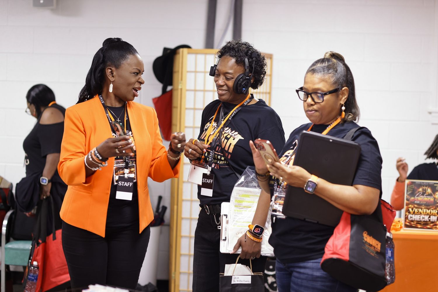 Three women with lanyards and name badges chat and smile while one holds a tablet at a busy indoor conference or networking event.