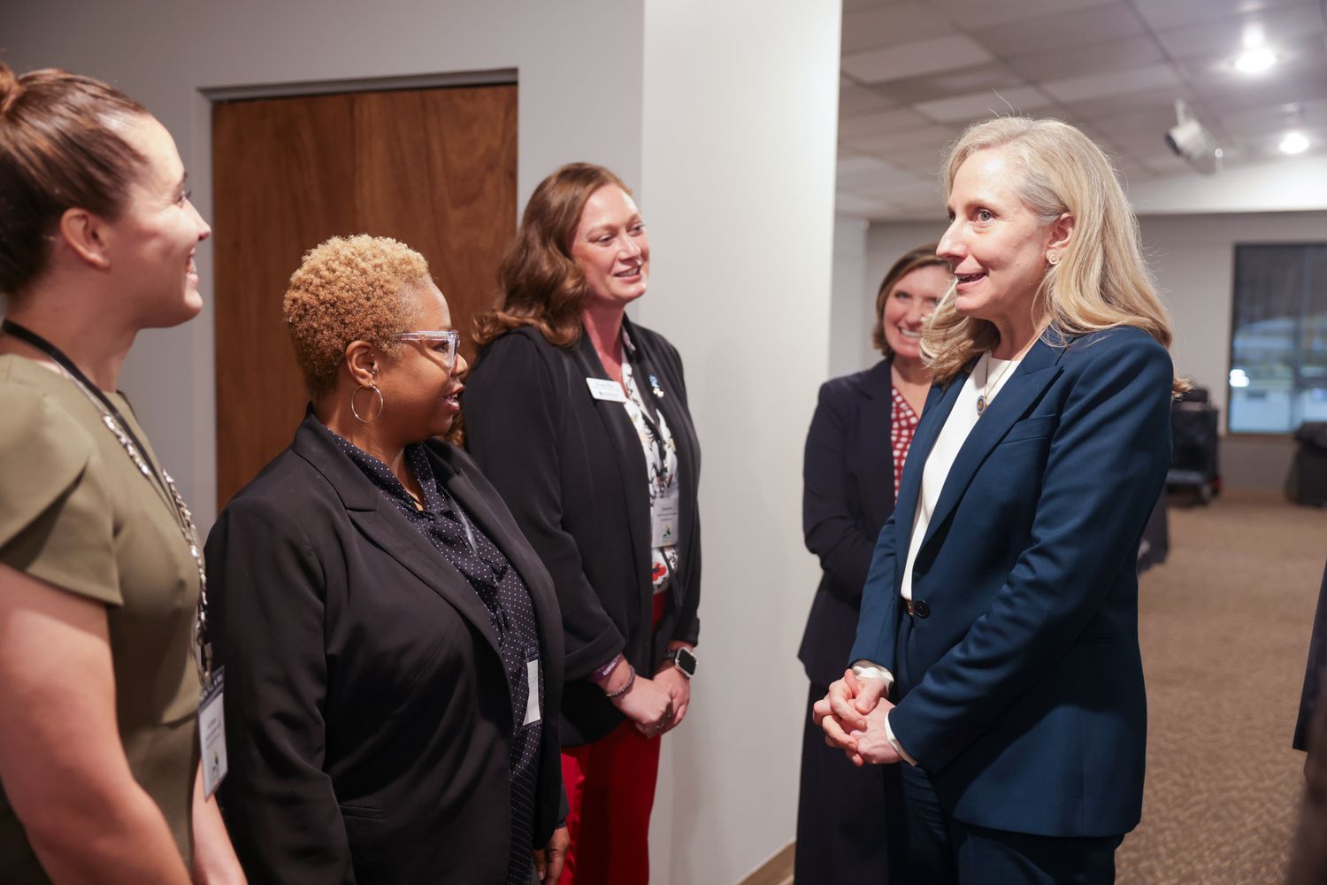 Group of professional women in business attire engaged in conversation in a hallway setting.