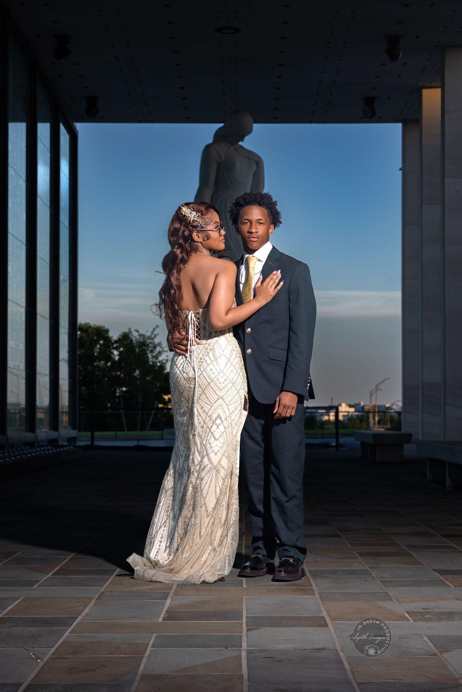 Elegant couple in formal prom attire posing at sunset, woman in beaded gown and man in navy suit with gold tie.