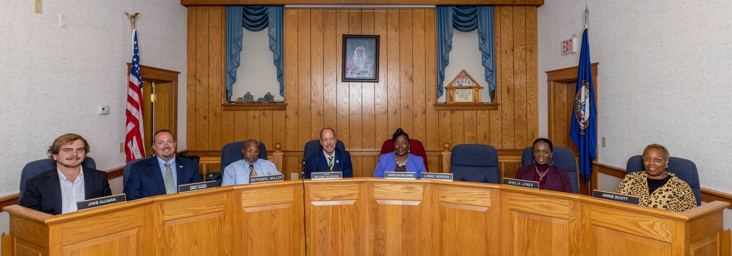 Seven council members seated at a wooden government chambers bench beneath American and Virginia state flags.