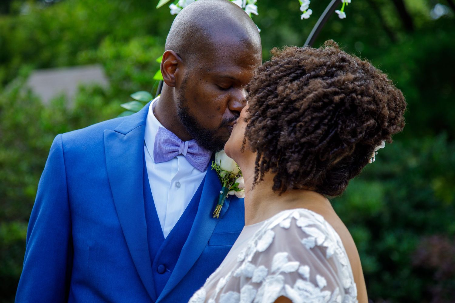 Bride and groom share a tender kiss outdoors surrounded by lush green garden foliage.