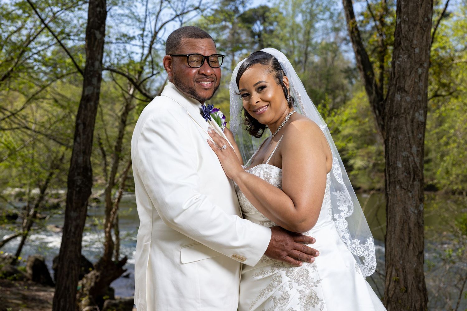 Couple in wedding attire embrace outdoors near a stream surrounded by spring trees.