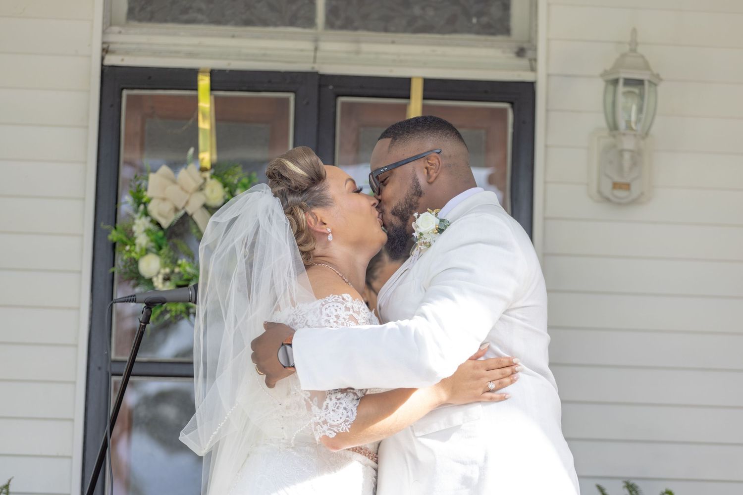 Bride and groom share a romantic kiss outside a white house with a floral wreath.
