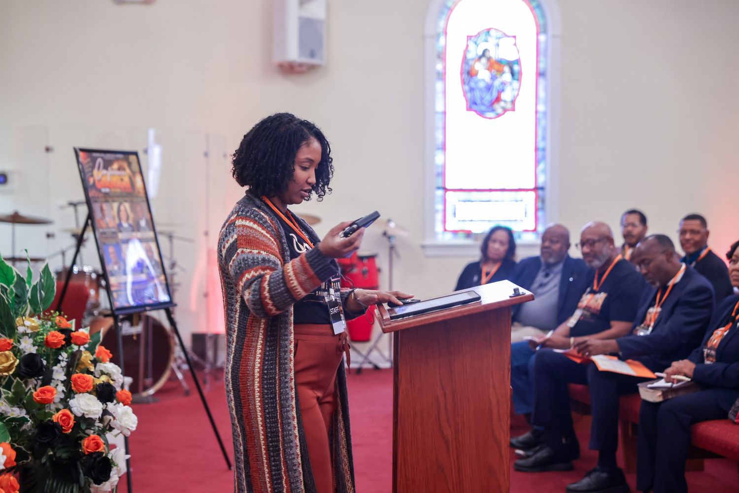 Woman in a striped cardigan speaks at a wooden podium in a church as seated attendees listen near a memorial photo and orange flowers.