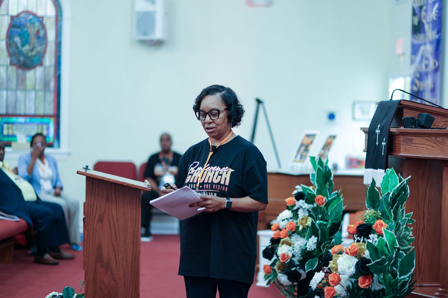 Woman in glasses and a black church t-shirt reads from papers at a wooden podium beside orange and white flowers during a church service.