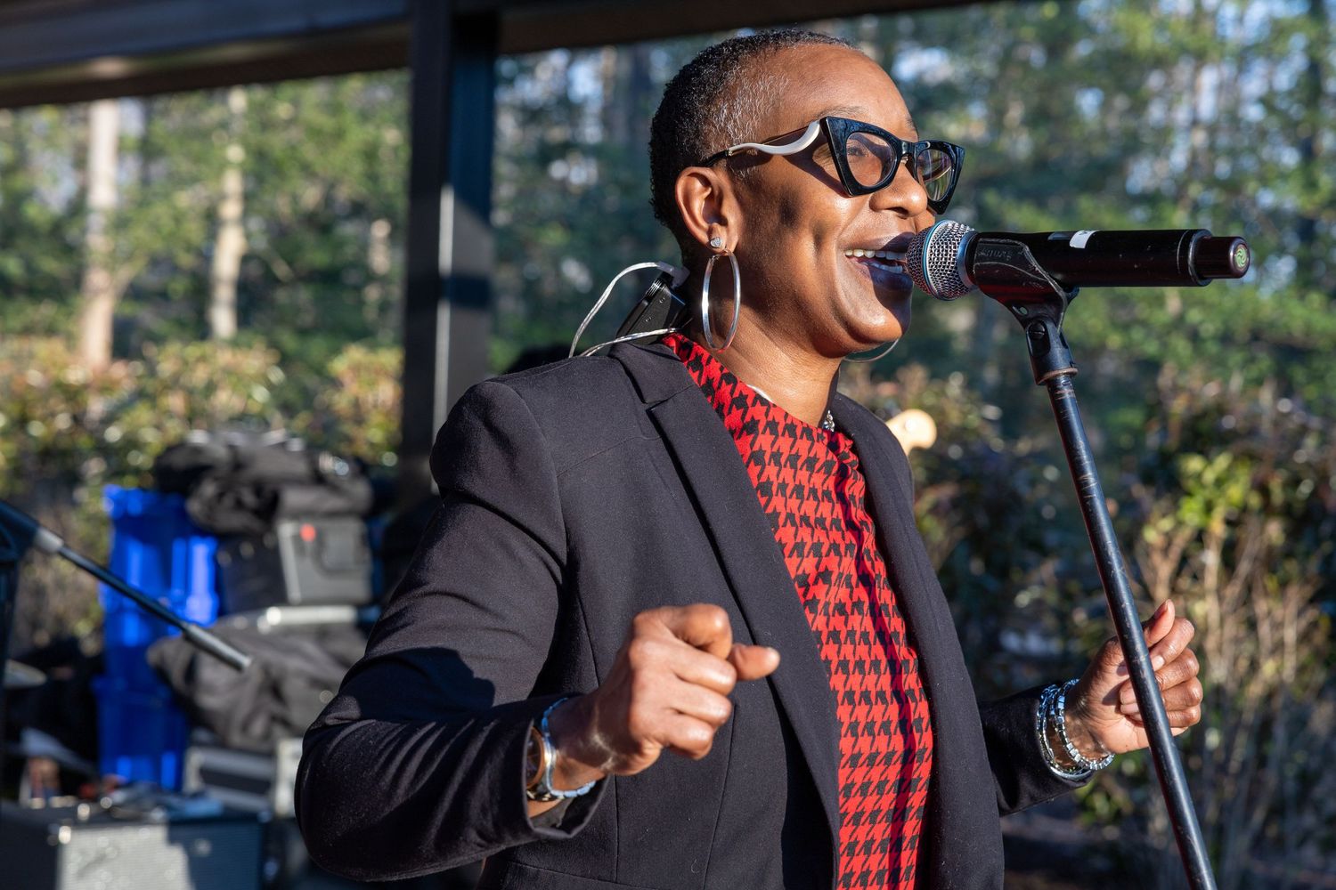 A performer sings passionately into a microphone wearing a black blazer and red houndstooth vest outdoors.