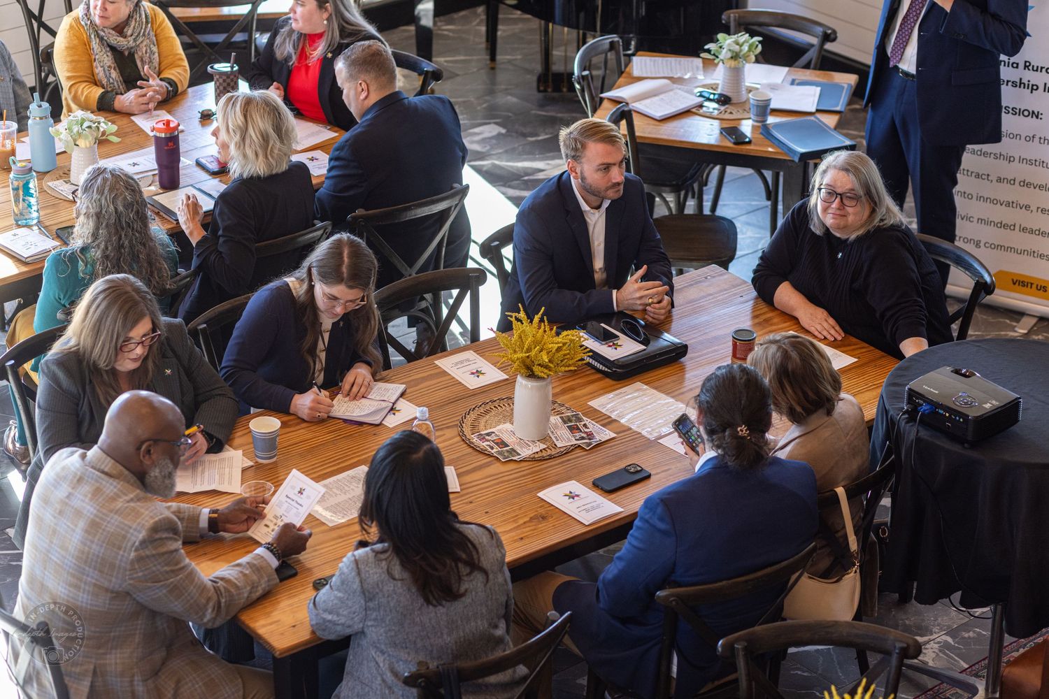 A diverse group of professionals engaged in discussion around wooden tables at an indoor networking event.