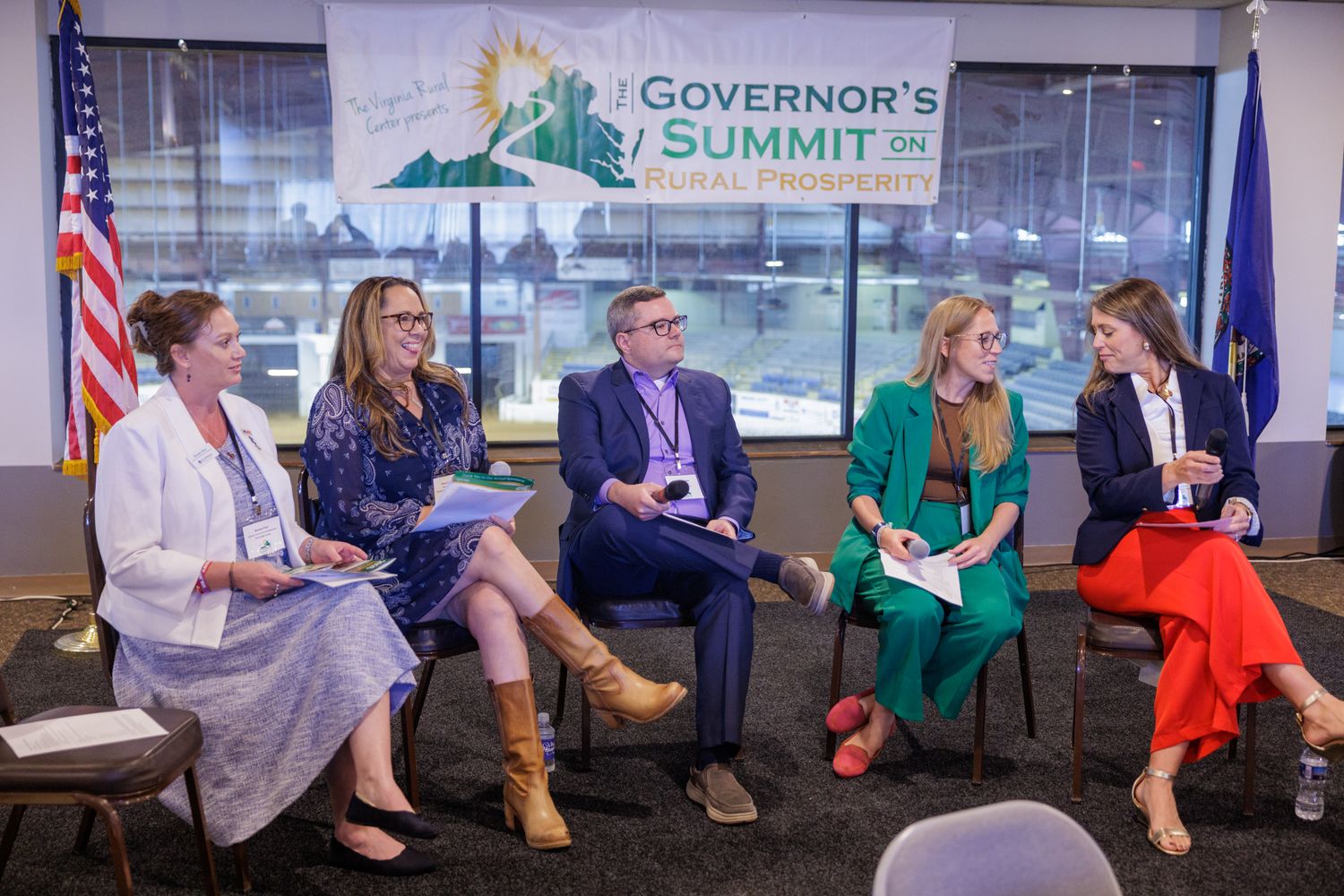 Six panelists seated in a semicircle at the Governor's Summit on Rural Prosperity event with US and state flags.