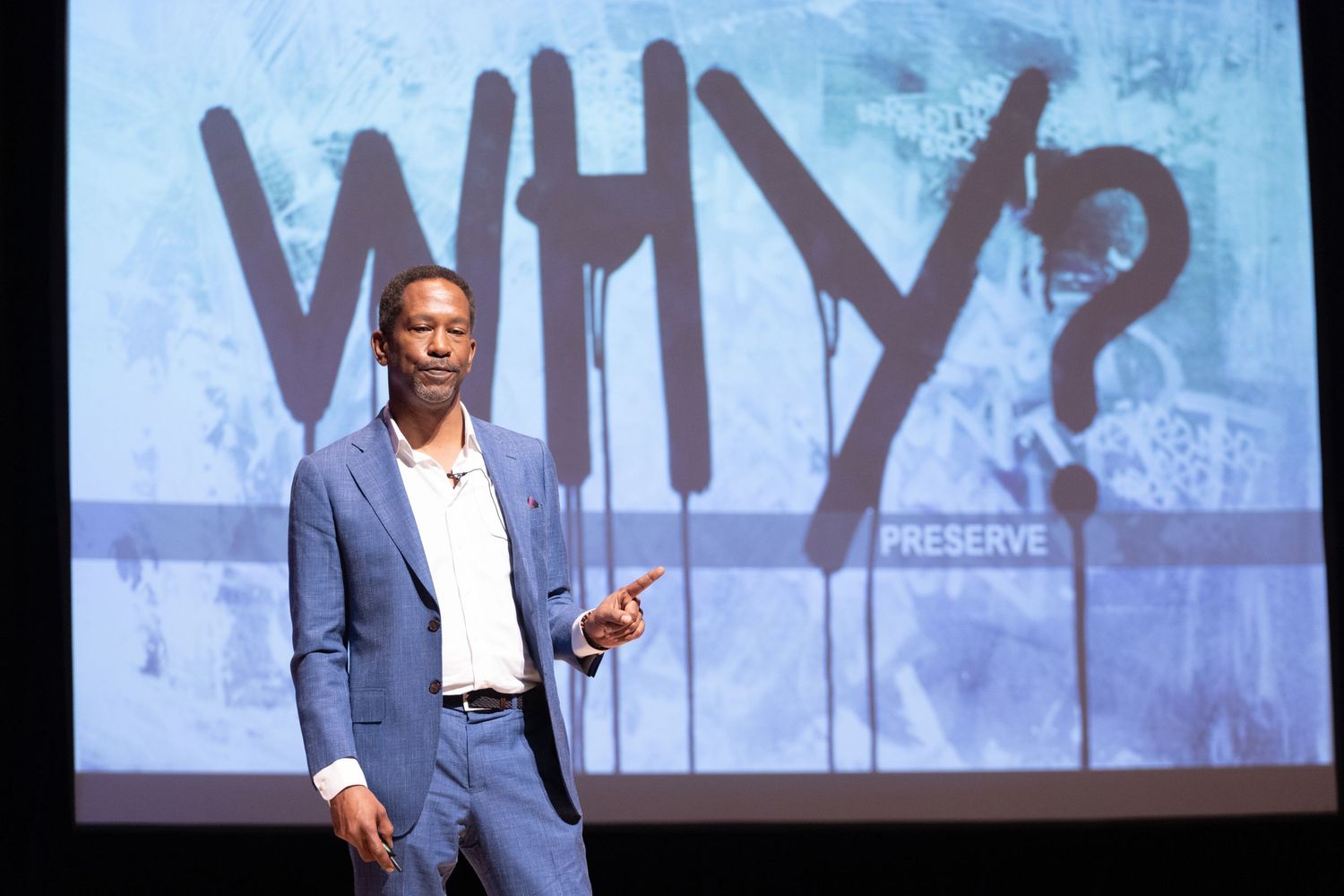 A speaker presents on stage in front of a large projection screen displaying the word WHY? at a preservation conference.