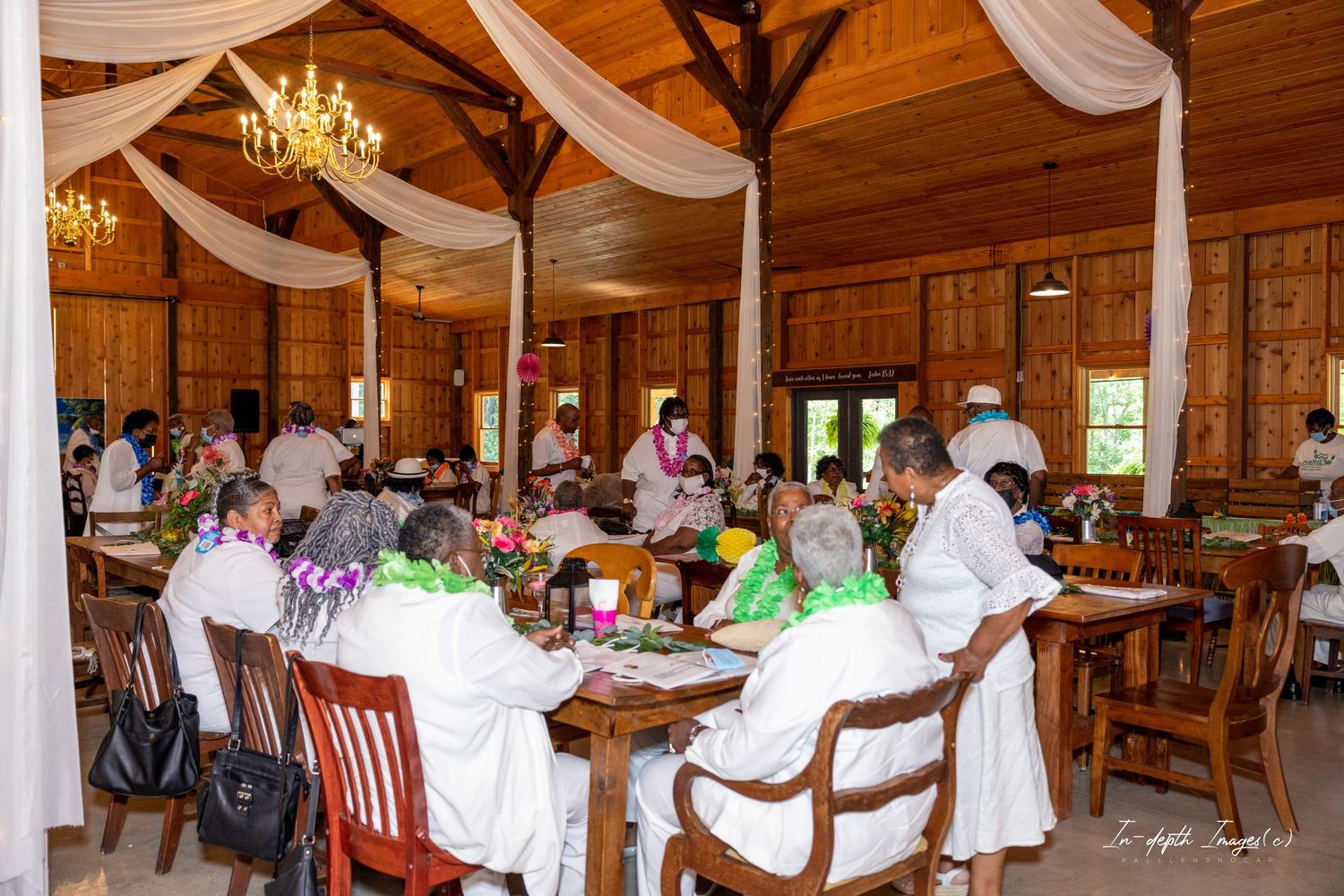 Guests wearing white attire and floral leis dine together in a decorated wooden barn venue with chandeliers.