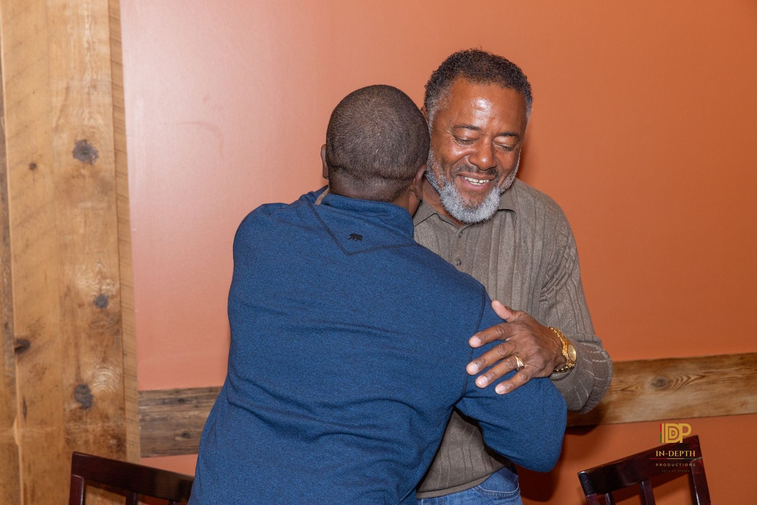 Two men share a warm embrace in a rustic restaurant, one smiling with a gray beard in a olive button-up shirt, the other in a blue shirt.