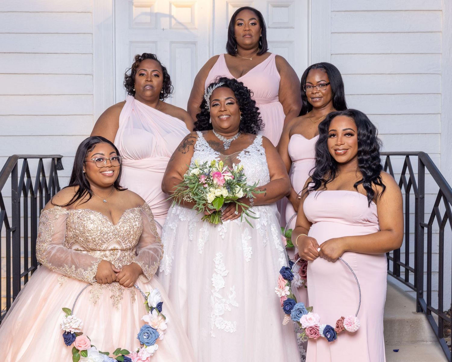 Bride poses with four bridesmaids in blush pink gowns on steps holding floral bouquets.