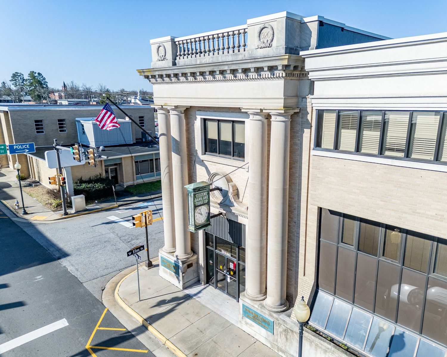 An aerial view of a neoclassical bank building with large columns, American flag, and surrounding intersection.