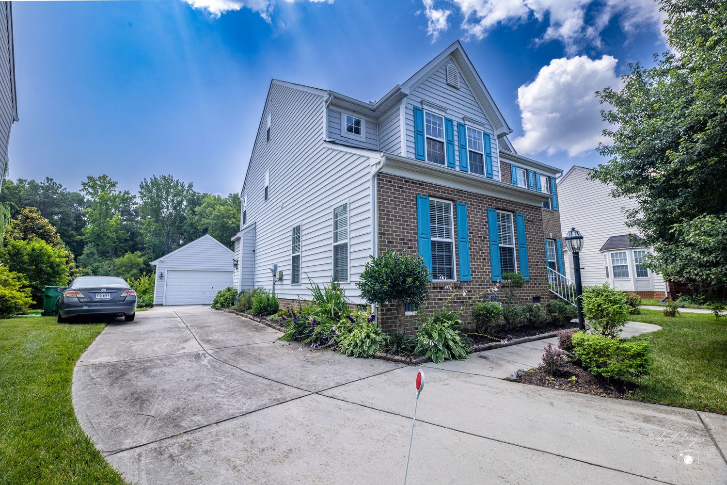 Two-story colonial home with brick and white siding, blue shutters, attached garage, and lush green landscaping on a sunny day.