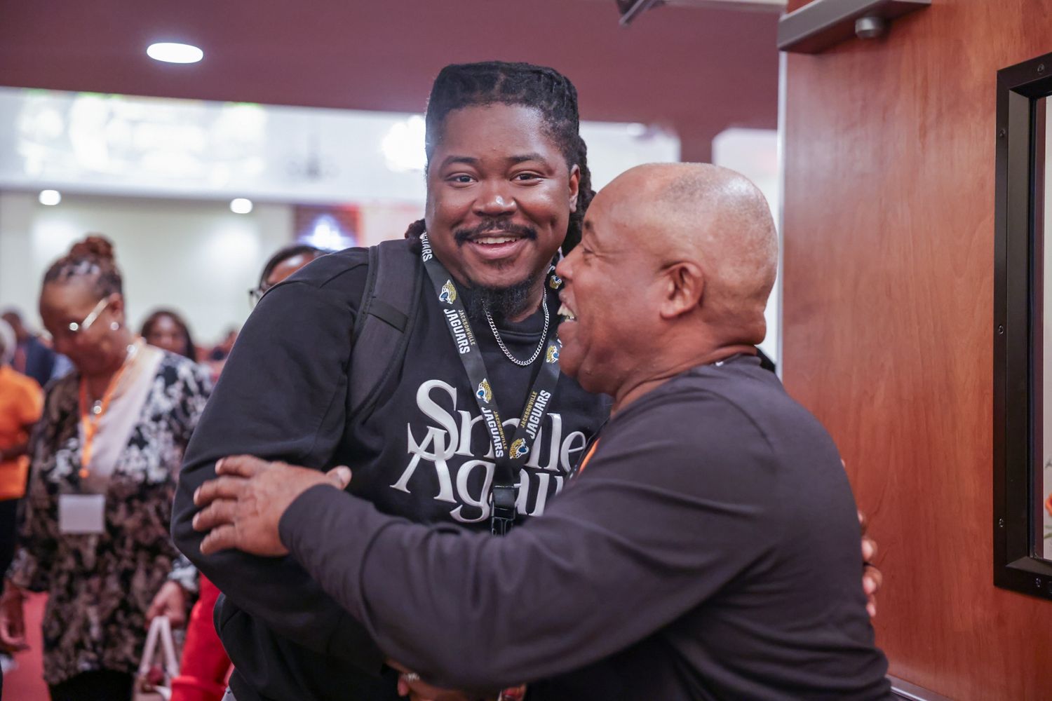 Two men share a warm, smiling embrace in a crowded indoor event, one wearing a lanyard and backpack with a graphic black shirt.