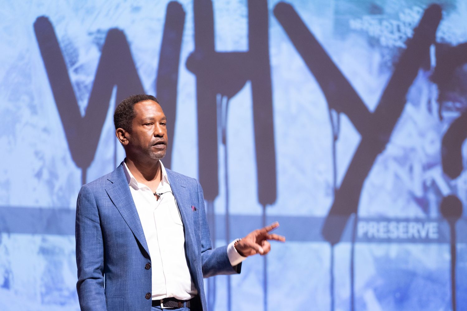 Man in blue blazer speaking on stage, pointing finger, with WHY PRESERVE projected on background screen.