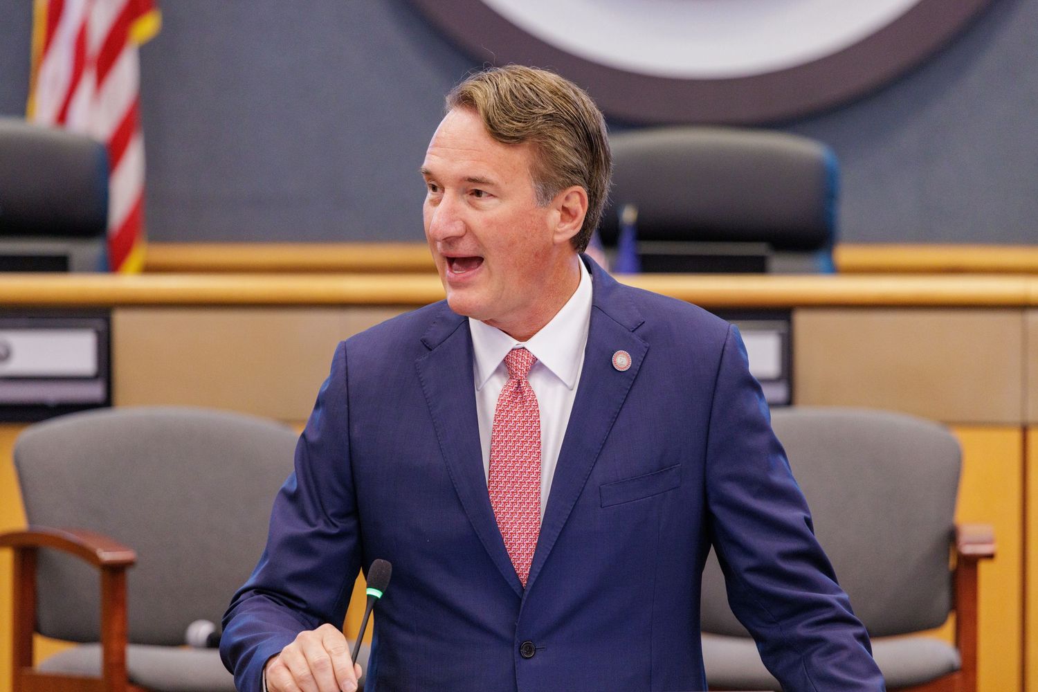 Man in navy blue suit with pink tie smiling in a formal government chamber setting.