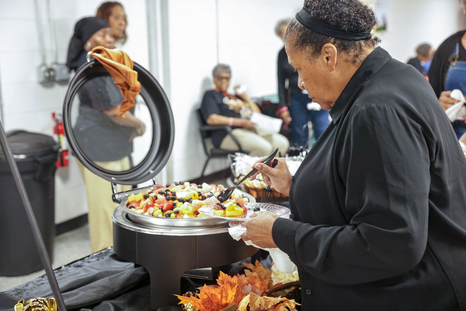 Woman in black serves fresh salad from a chafing dish at a church fellowship meal while guests sit and chat in the background.