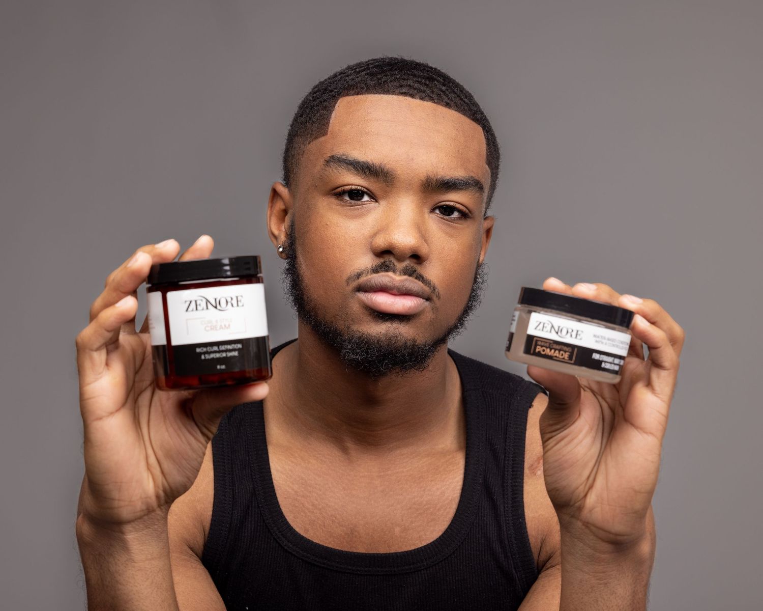 Young Black man in black tank top holding two Zenore grooming product jars with a serious expression.