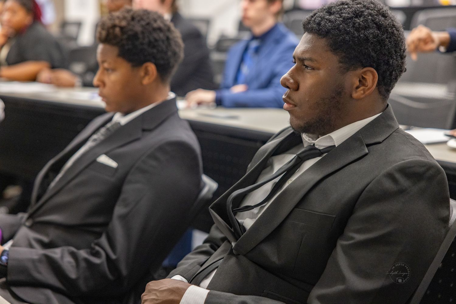 Two young men in suits sit attentively in a professional meeting or classroom setting.