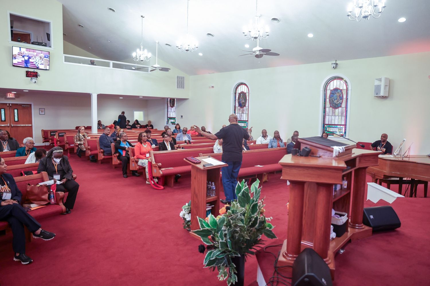 Congregation seated in wooden pews of a church sanctuary with red carpet, stained glass windows, and a man standing near the pulpit.