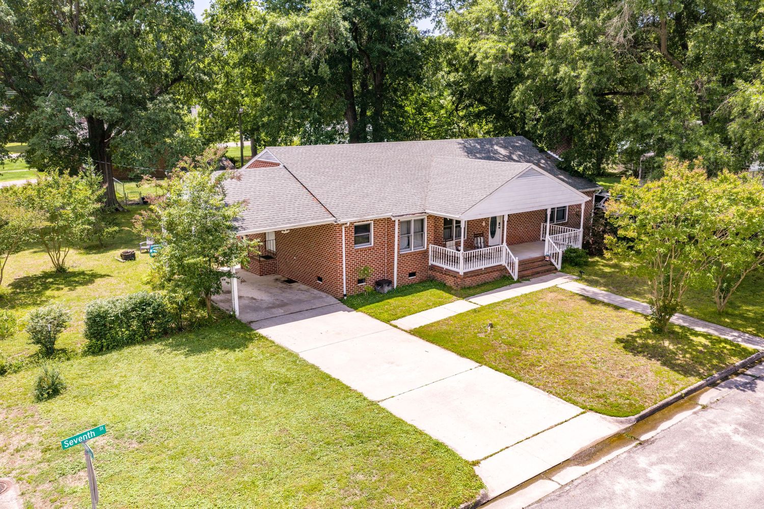 Aerial view of a brick ranch home with a covered porch, attached carport, concrete driveway, and lush green lawn surrounded by trees.