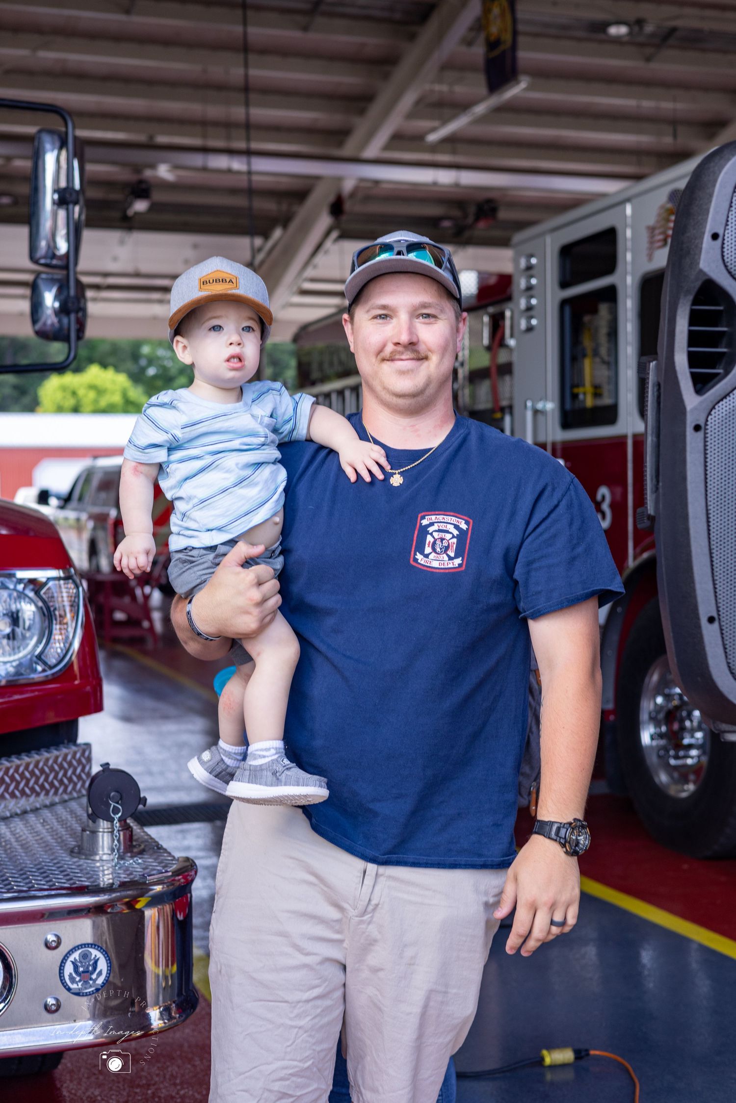 Smiling firefighter holding young toddler in front of red fire truck at station.