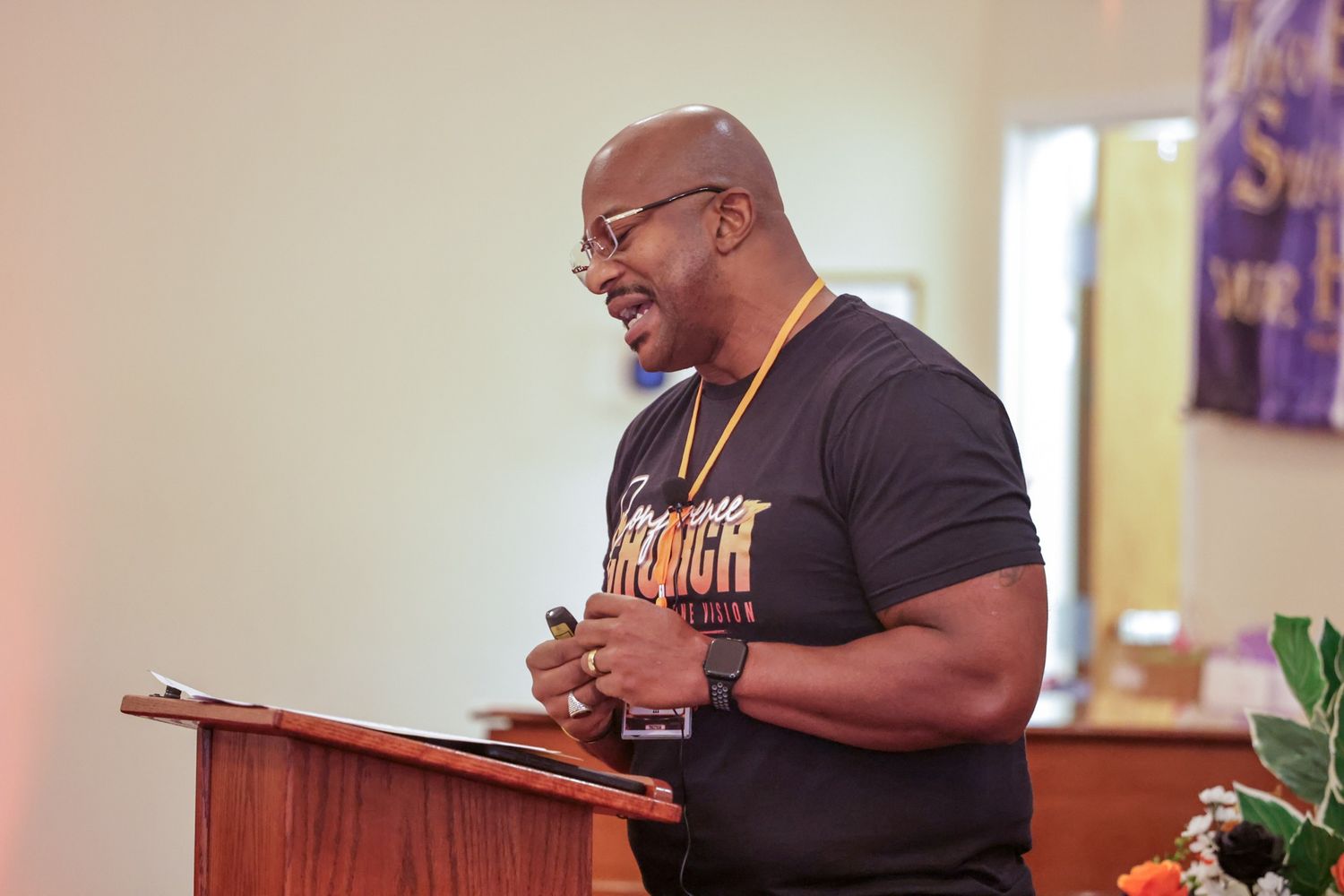 Man with glasses and a lanyard speaks from a wooden podium in a church sanctuary, wearing a black graphic t-shirt and holding a microphone.