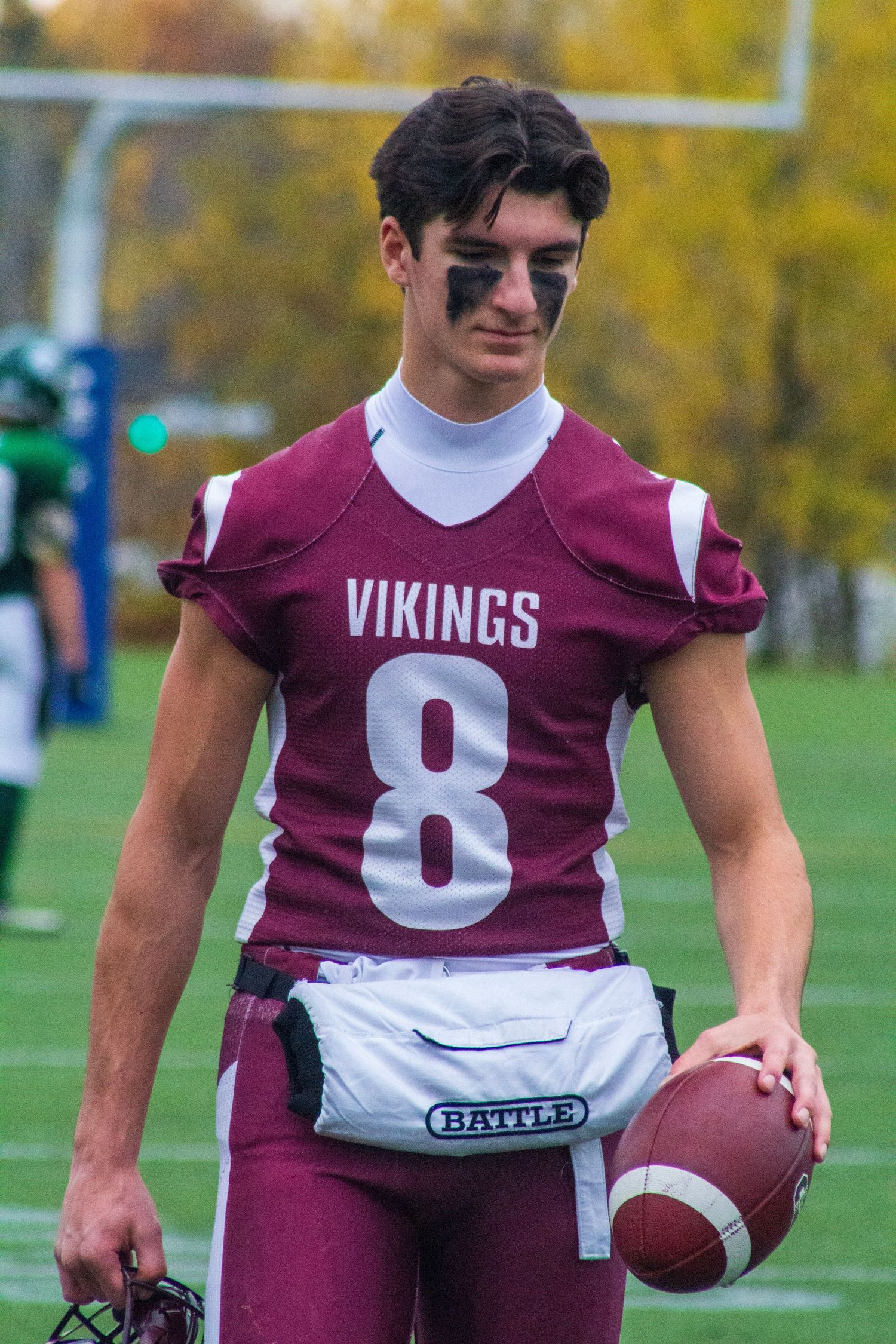 Young male football player in Vikings #8 maroon jersey holding helmet and football on green field.