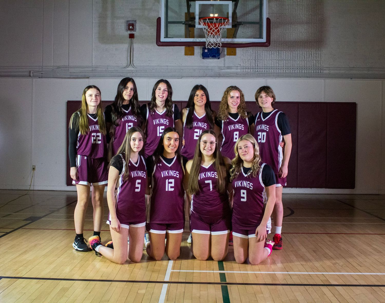 Girls youth basketball team in maroon Vikings uniforms posing together on indoor gym court for team photo.