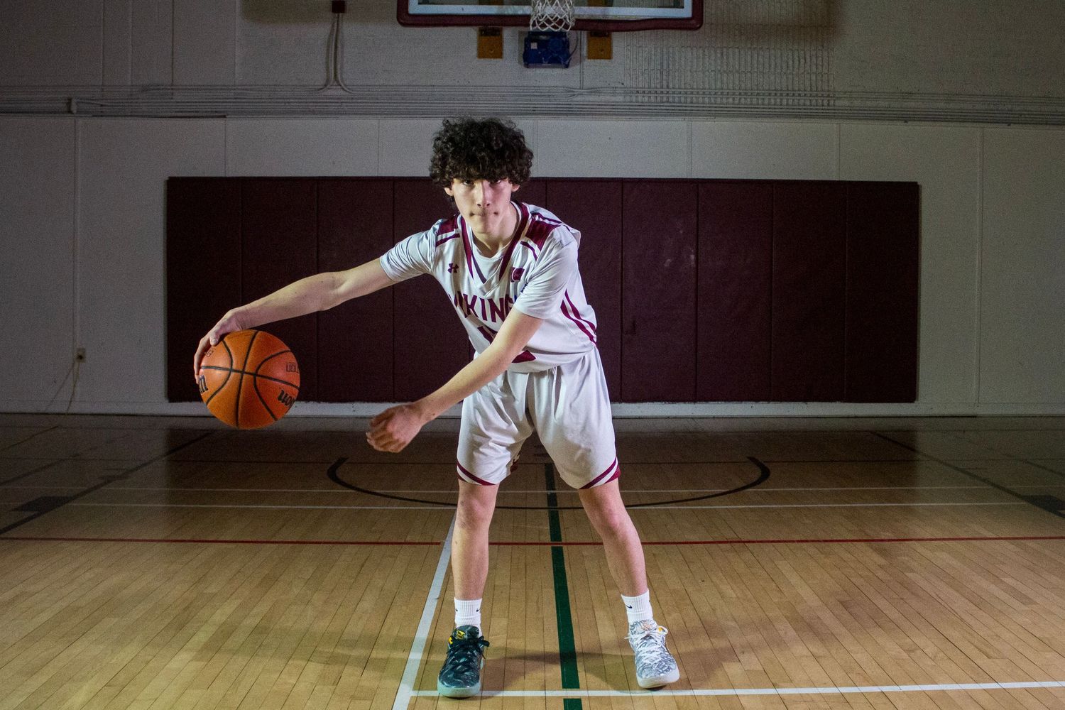 Young basketball player dribbling on indoor court wearing white and maroon uniform.