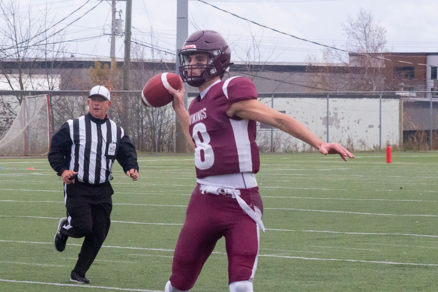 Football player wearing number 8 maroon jersey throws a pass during a game with a referee watching nearby.