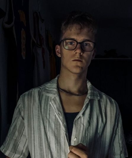 Young man with glasses and chain necklace wearing striped shirt in dramatic dark lighting portrait.