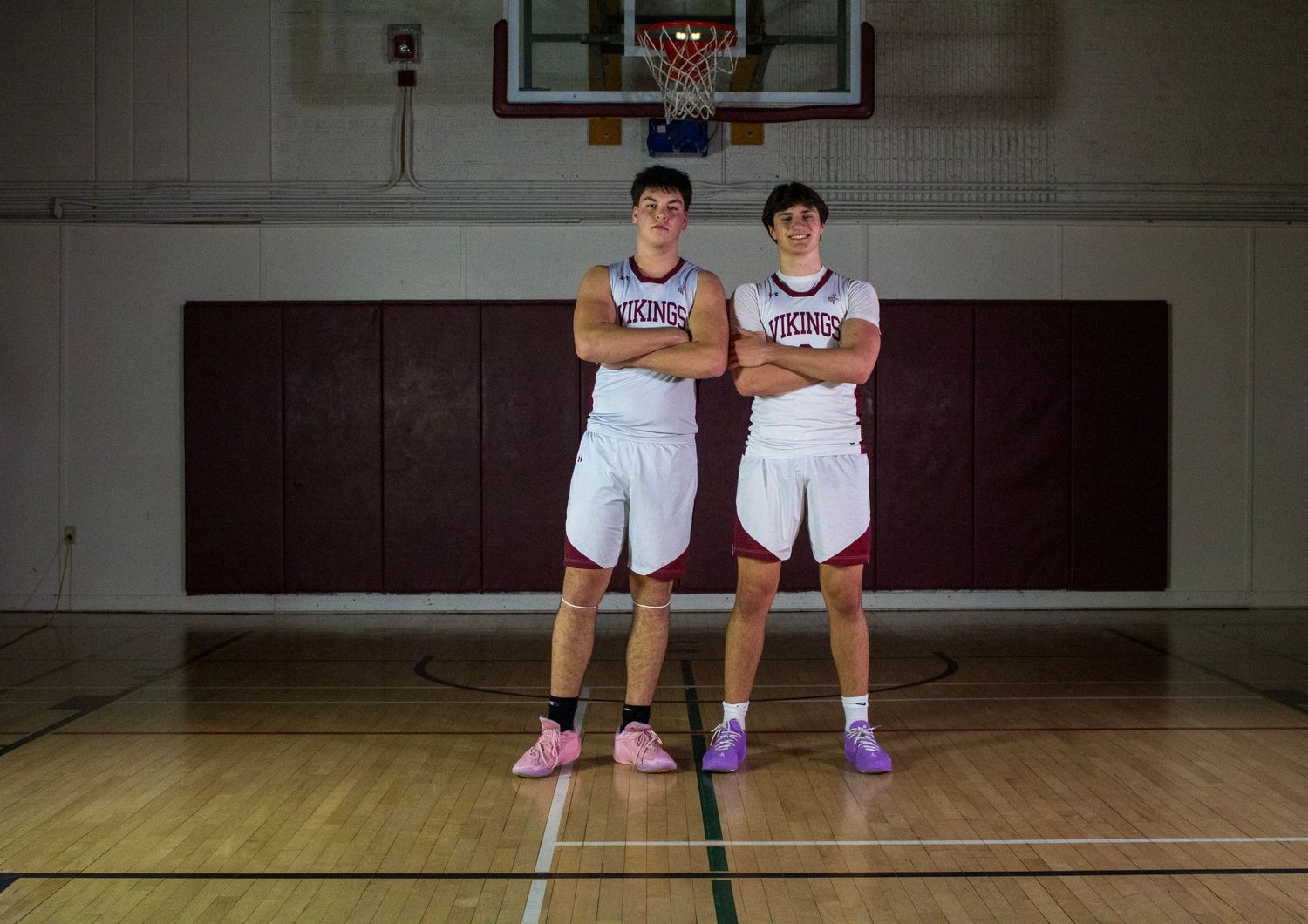 Two high school basketball players in white uniforms pose with arms crossed on an indoor court.