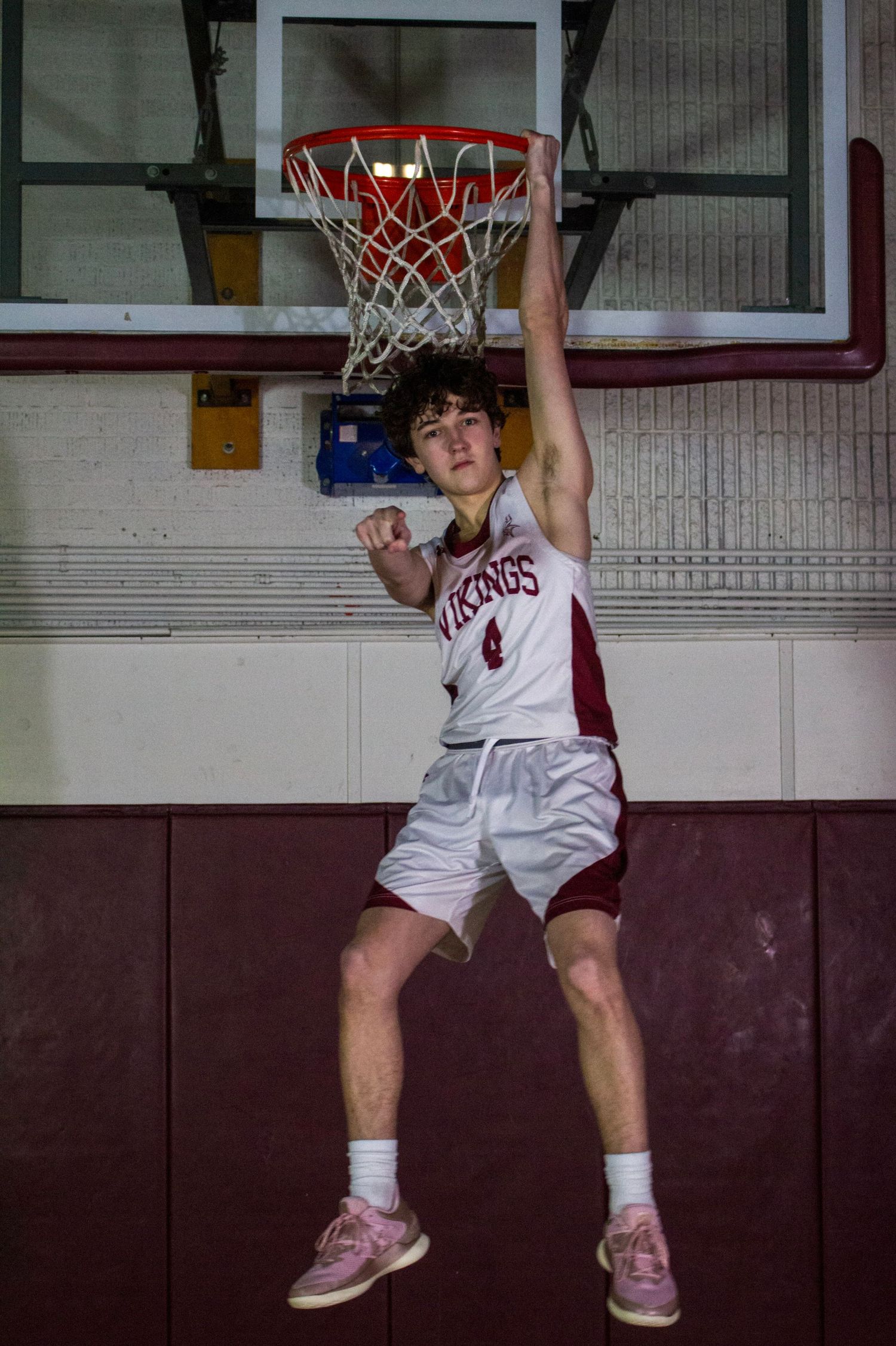 Young basketball player in Vikings jersey hanging from hoop in gym.