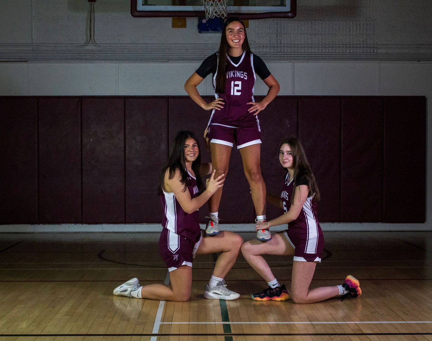 Three female Vikings basketball athletes pose together in a gym, wearing maroon uniforms.
