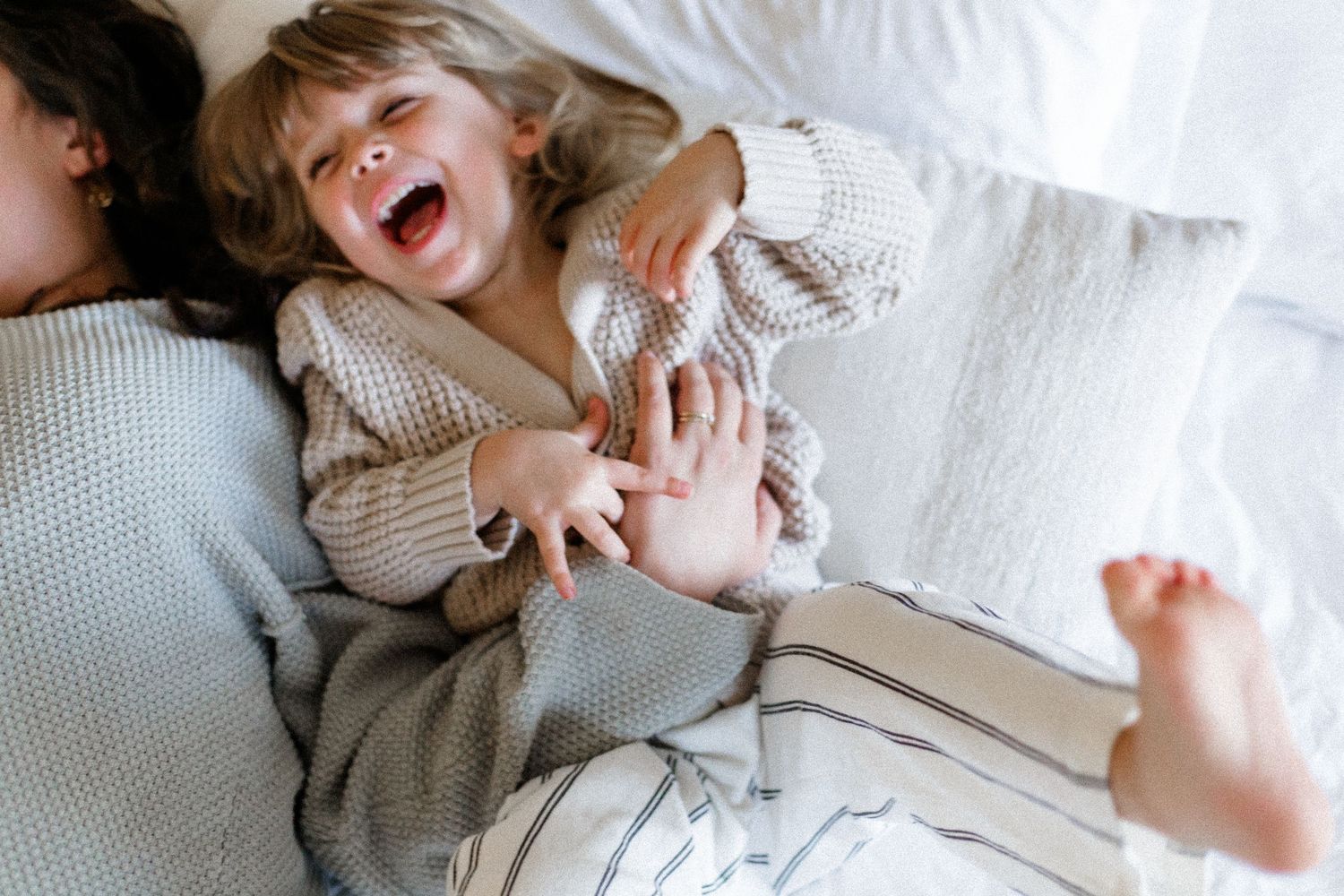 A child in a knit sweater laughs joyfully while lying on white bedding.