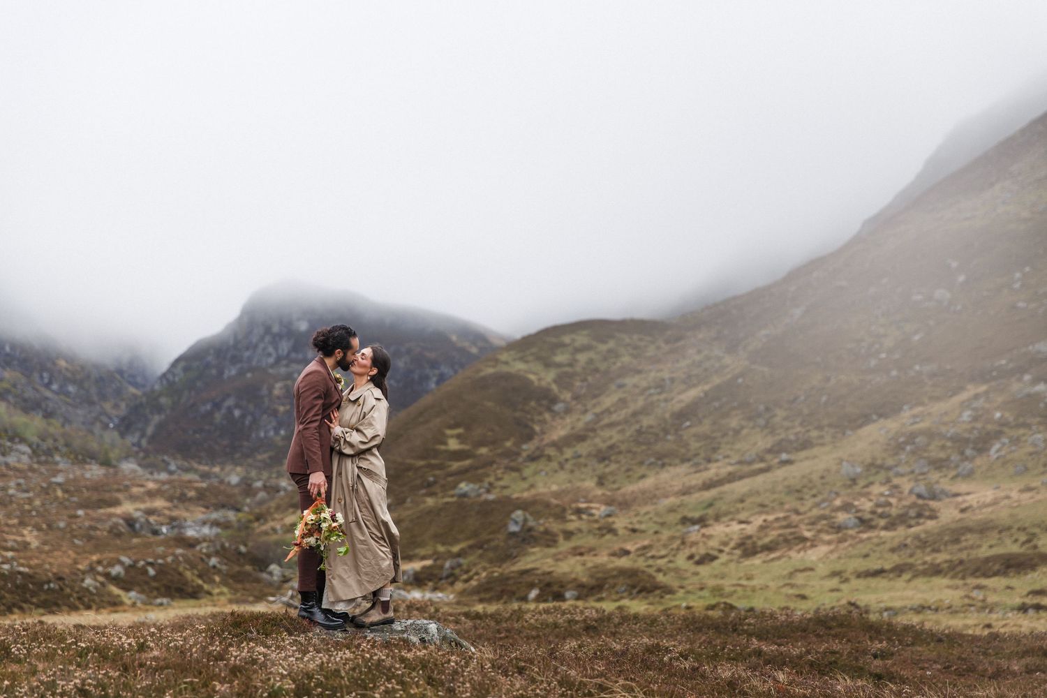 A couple embraces in the misty Scottish Highlands with rugged mountains and rolling terrain in the background.
