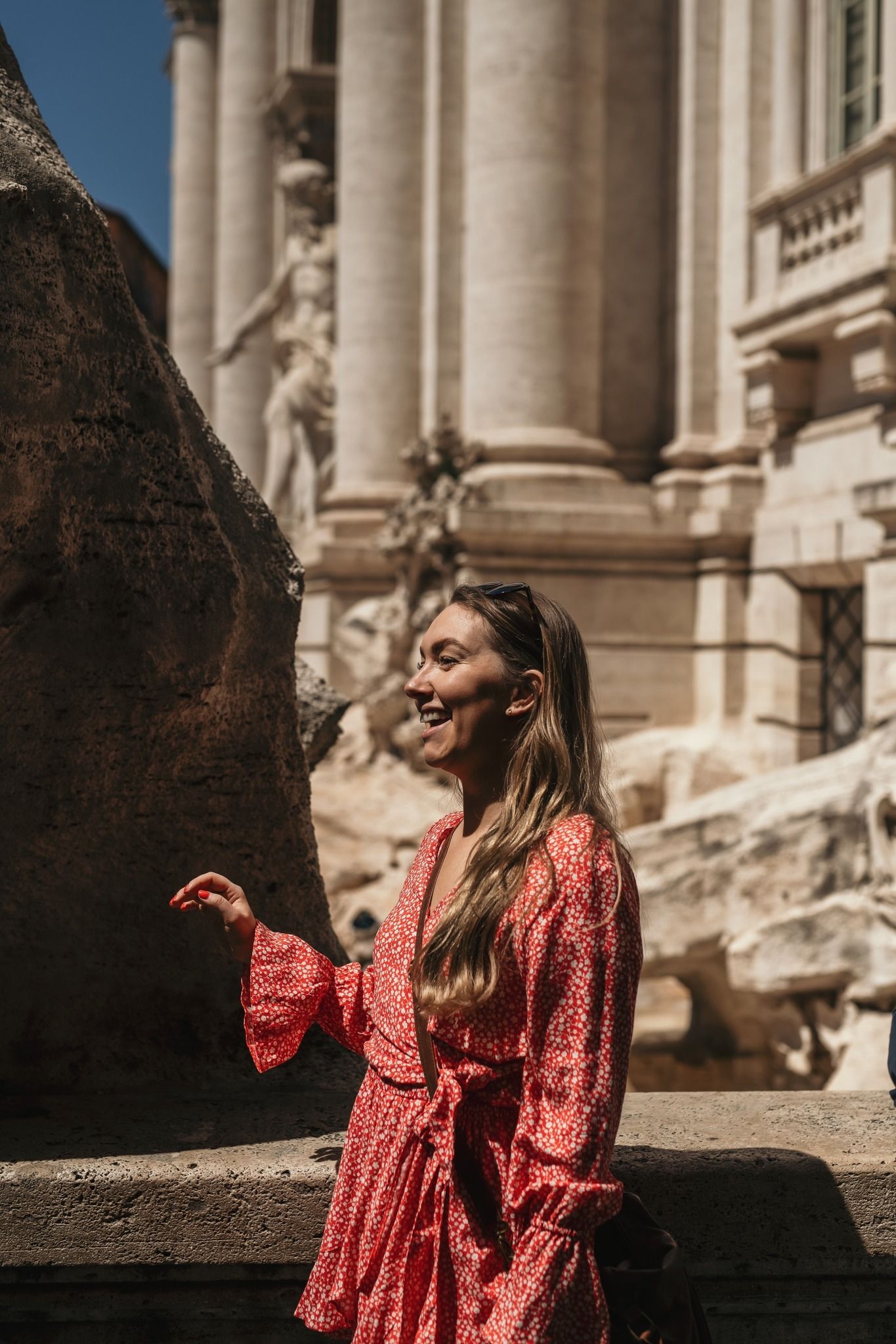 Woman in flowy red polka dot dress stands by ornate classical architecture with stone columns and sculptures.