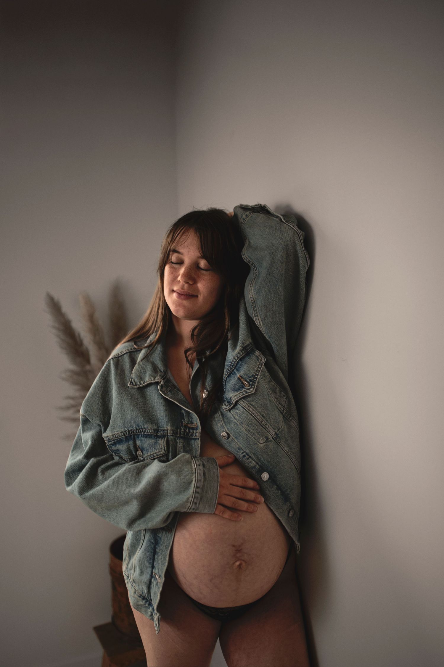 A pregnant woman wearing an oversized denim jacket poses against a wall with pampas grass decoration.