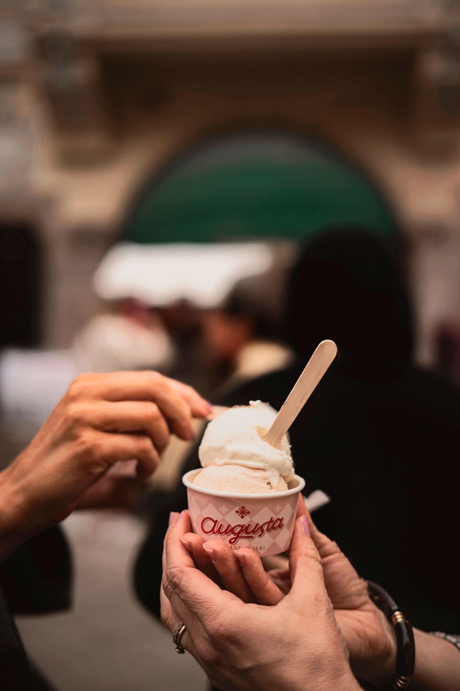 Hands holding Angermeier branded ice cream cup with wooden spoon and creamy dessert against blurred background.
