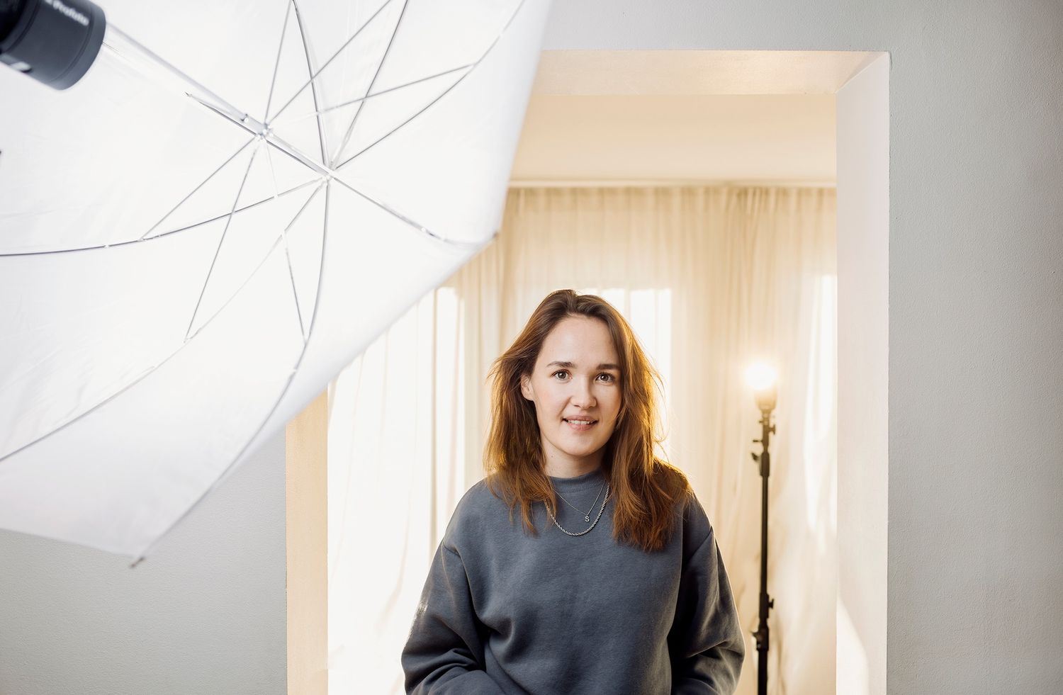Person in gray sweater stands in doorway with photography lighting umbrella equipment visible in foreground.