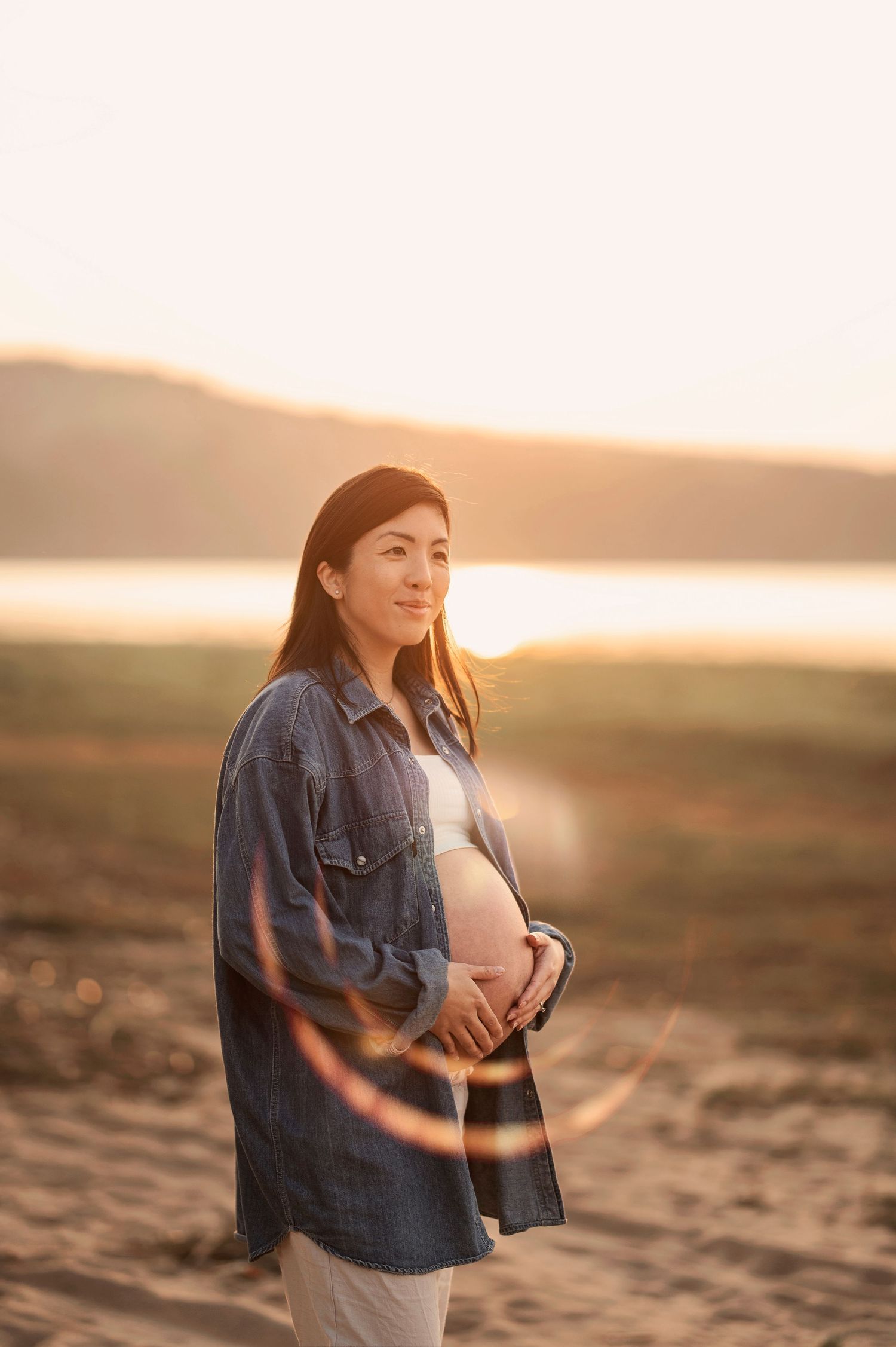 An expectant mother in denim jacket stands on beach at sunset, cradling her baby bump in warm golden light.