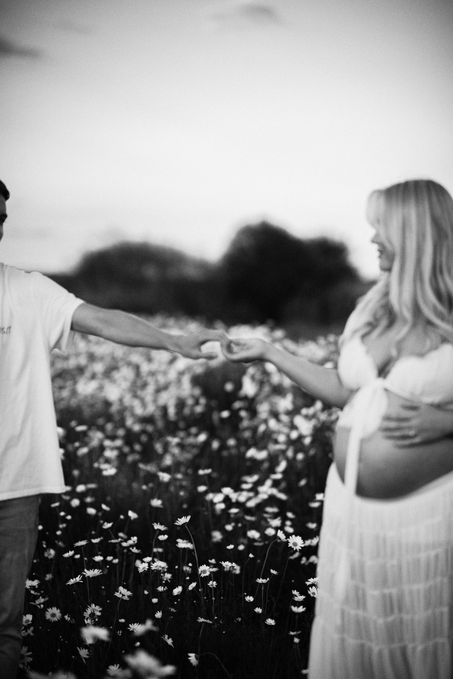 Black and white romantic photo of a couple holding hands in a field of wildflowers with a pregnant woman in a white dress.