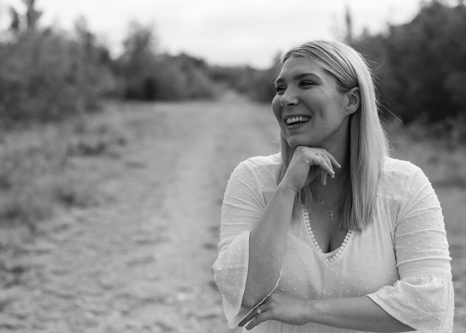 Black and white photo of a smiling blonde woman on a dirt road outdoors.