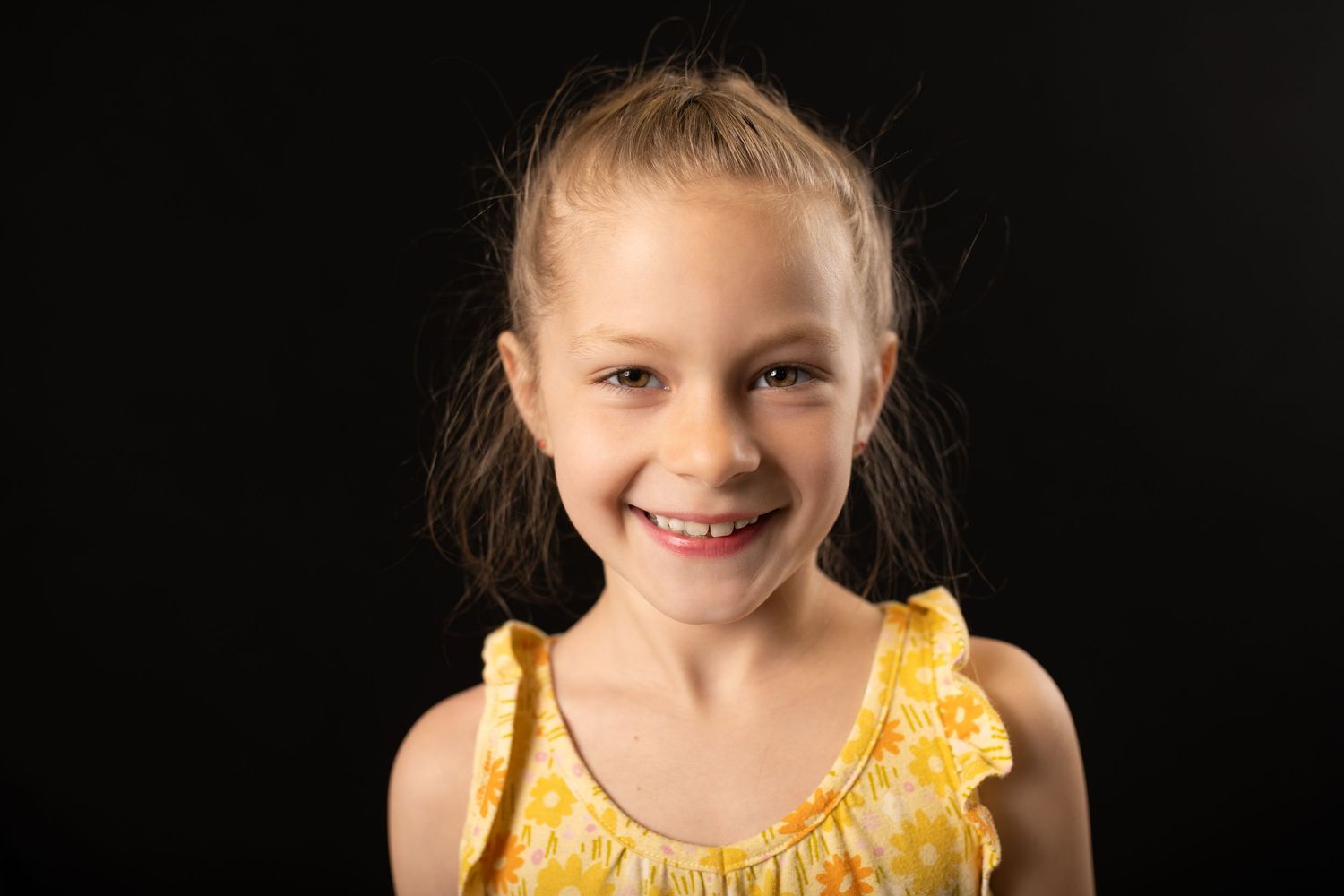 Cheerful young girl in a yellow dress smiling brightly against a dark background.