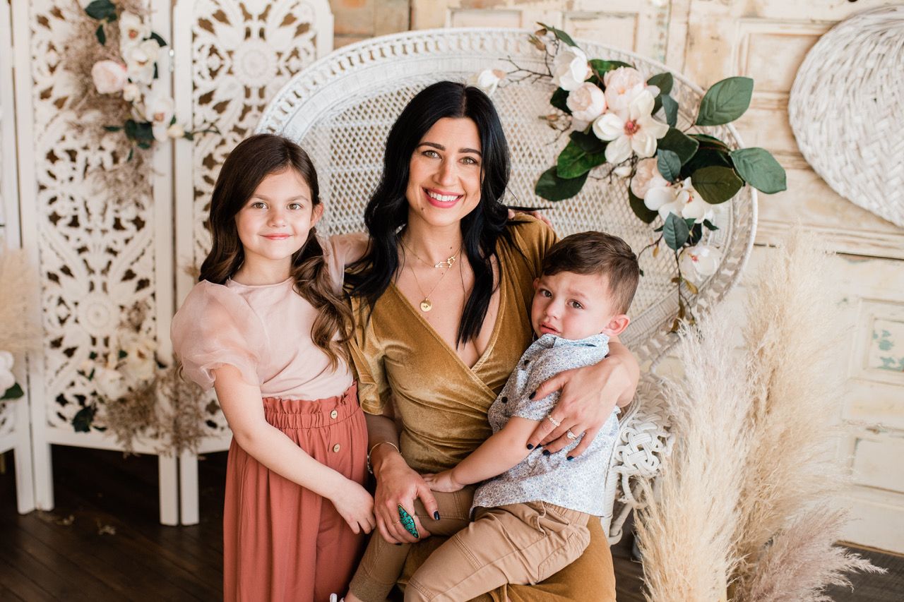 A smiling mother in a gold dress poses with her two young children in a boho-styled studio.