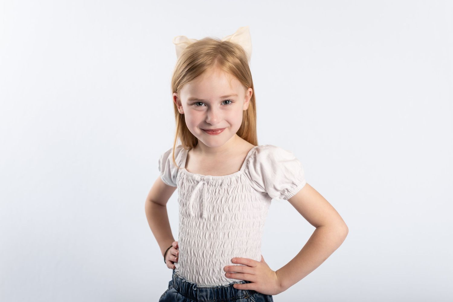 Young blonde girl wearing cat ears headband smiling with hands on hips.